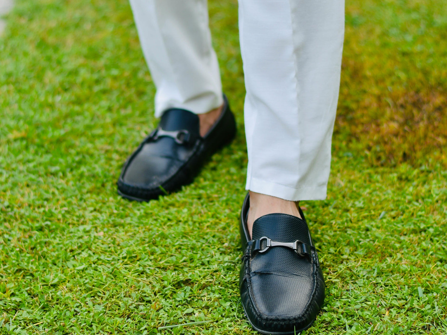 Person wearing black loafers with no-show socks and white pants, standing on green grass.