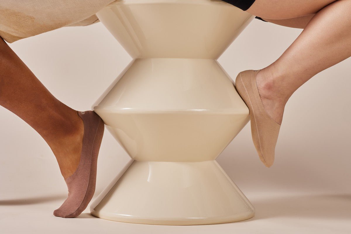 Two feet wearing no-show socks, one in brown and the other in beige, resting on opposite sides of a geometric cream-colored stool.